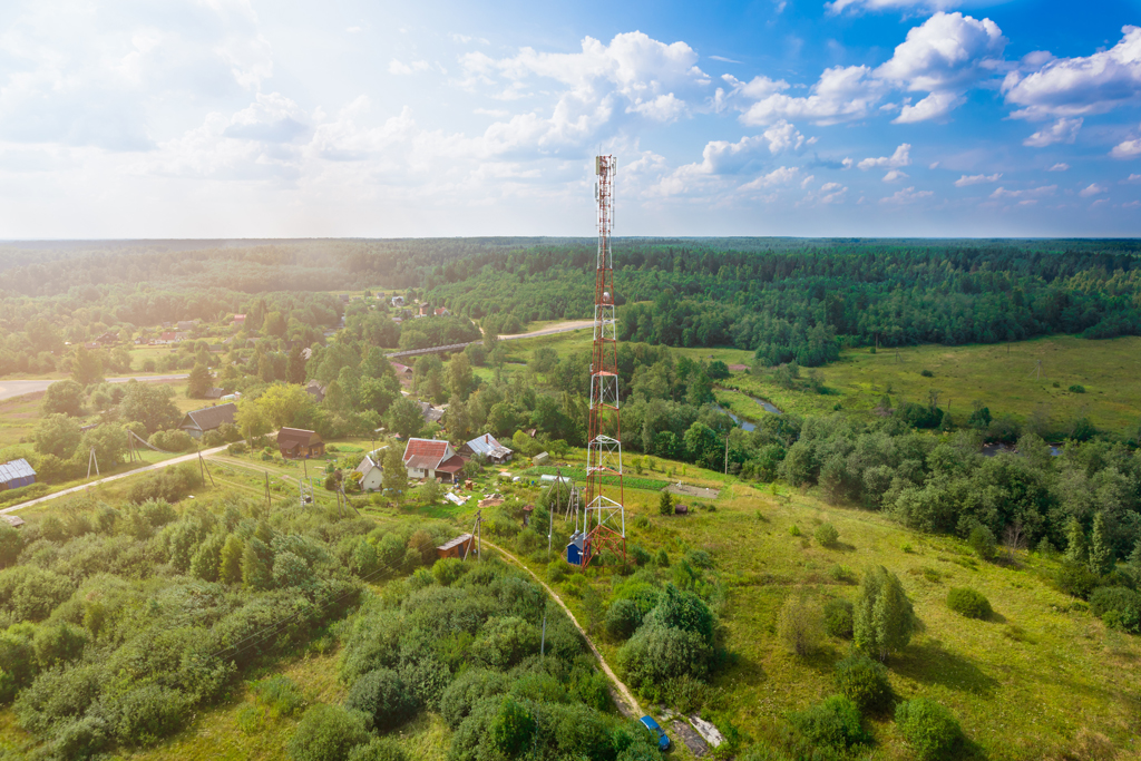 Telecommunication tower with radio antennas and satellite dishes is installed on the rural on the green field with grass, bushes and trees. Concept of harmless of electromagnetic and microwave radiation on the environment, nature and human. Telecommunication tower with radio antennas