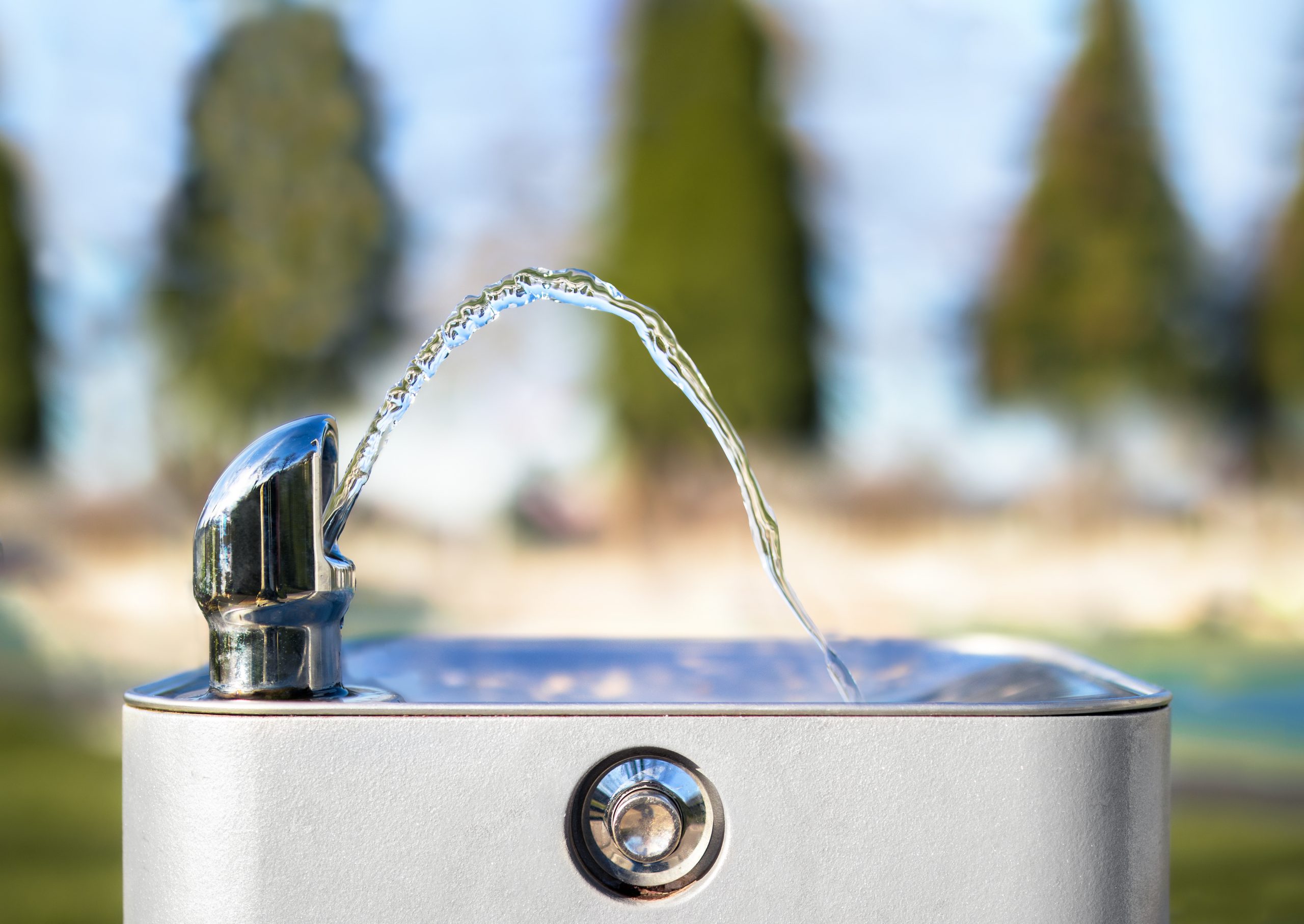 Drinking water fountain in park on a sunny day, no person. Close up of flowing water from the tab in an arch. Bright defocused park and tree background. Selective focus.