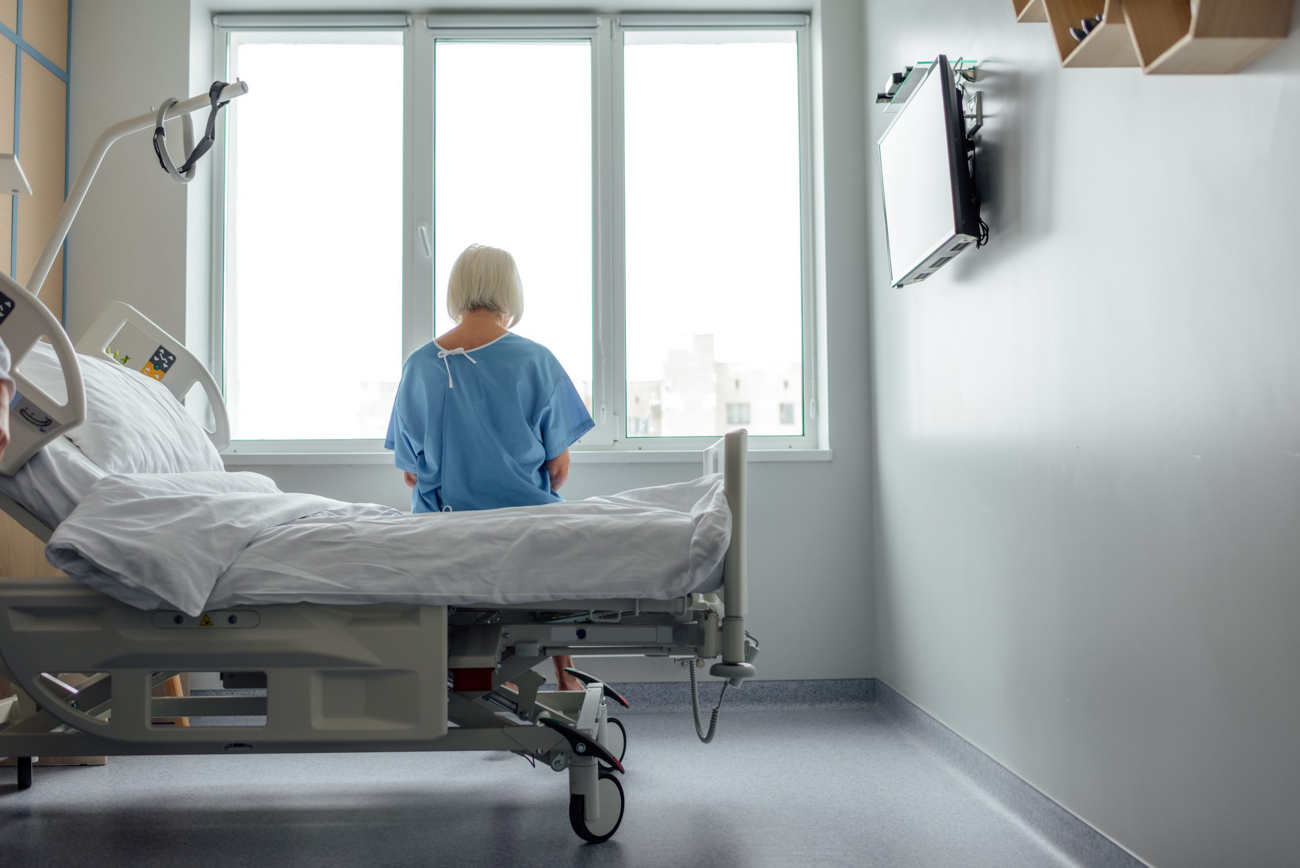 back view of lonely senior woman sitting on bed in hospital ward with copy space